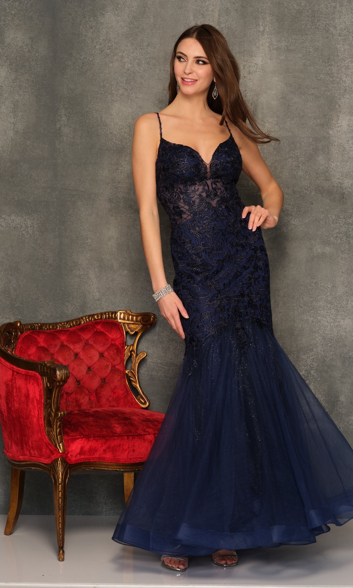 A woman wearing the Dave & Johnny A10625 Long Lace Mermaid Prom Dress stands beside an ornate red and gold armchair against a gray textured backdrop.